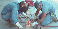 Two workers cutting and welding metal rods. Workers can be seen wearing gloves and eye protection. By National Institute for Occupational Safety and Health.